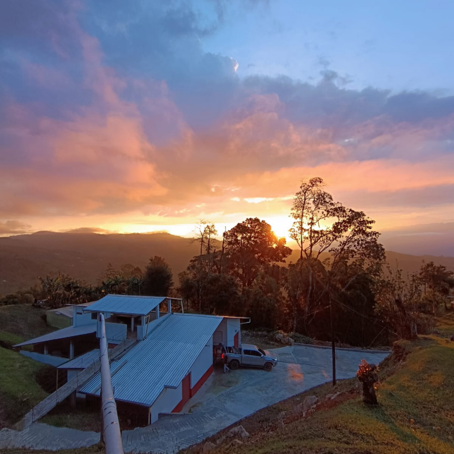 The sun peaks through pink and blue clouds and lights up trees and mountains behind a coffee farm house.