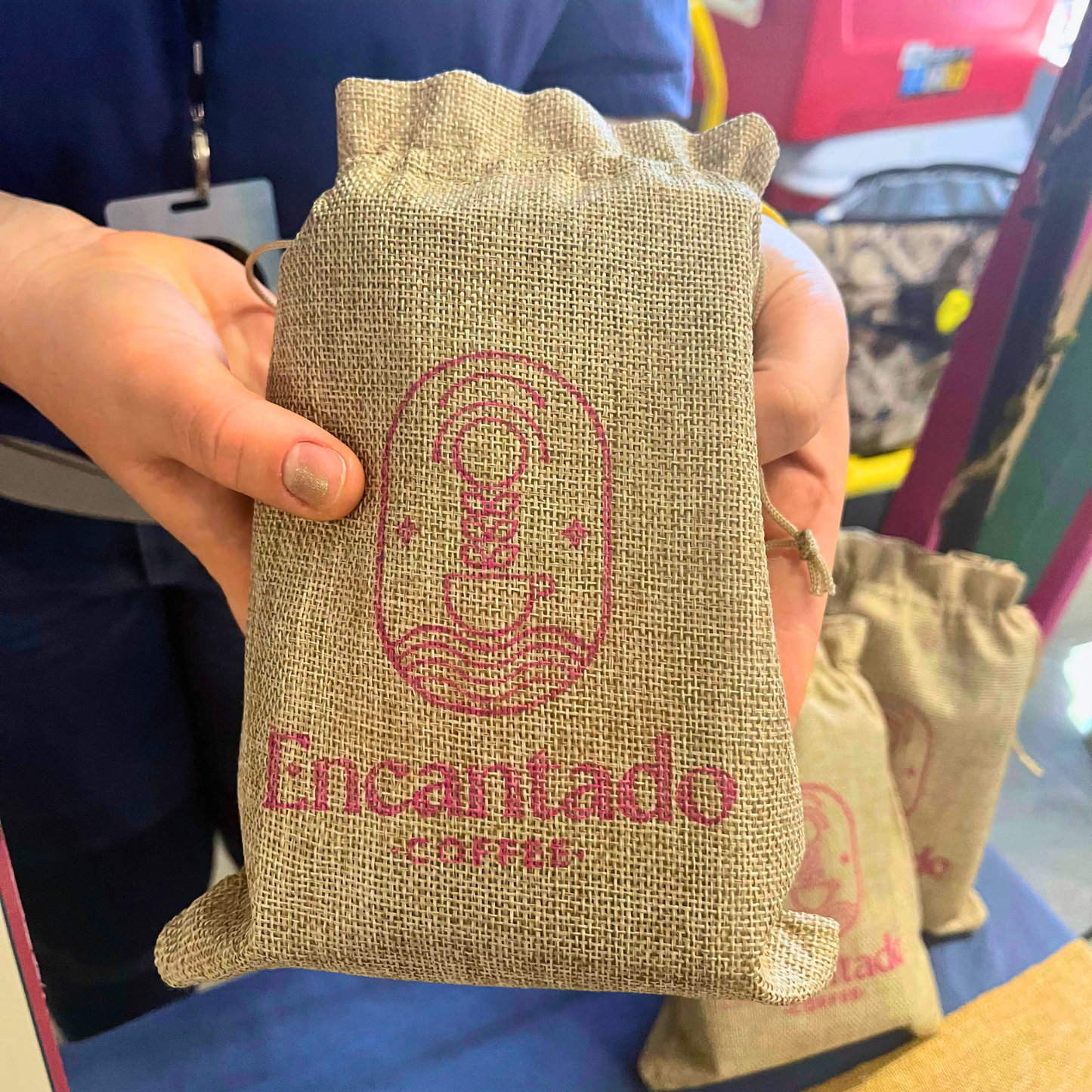 A hand holds a burlap bag with Encantado Coffee's logo over a blue table.