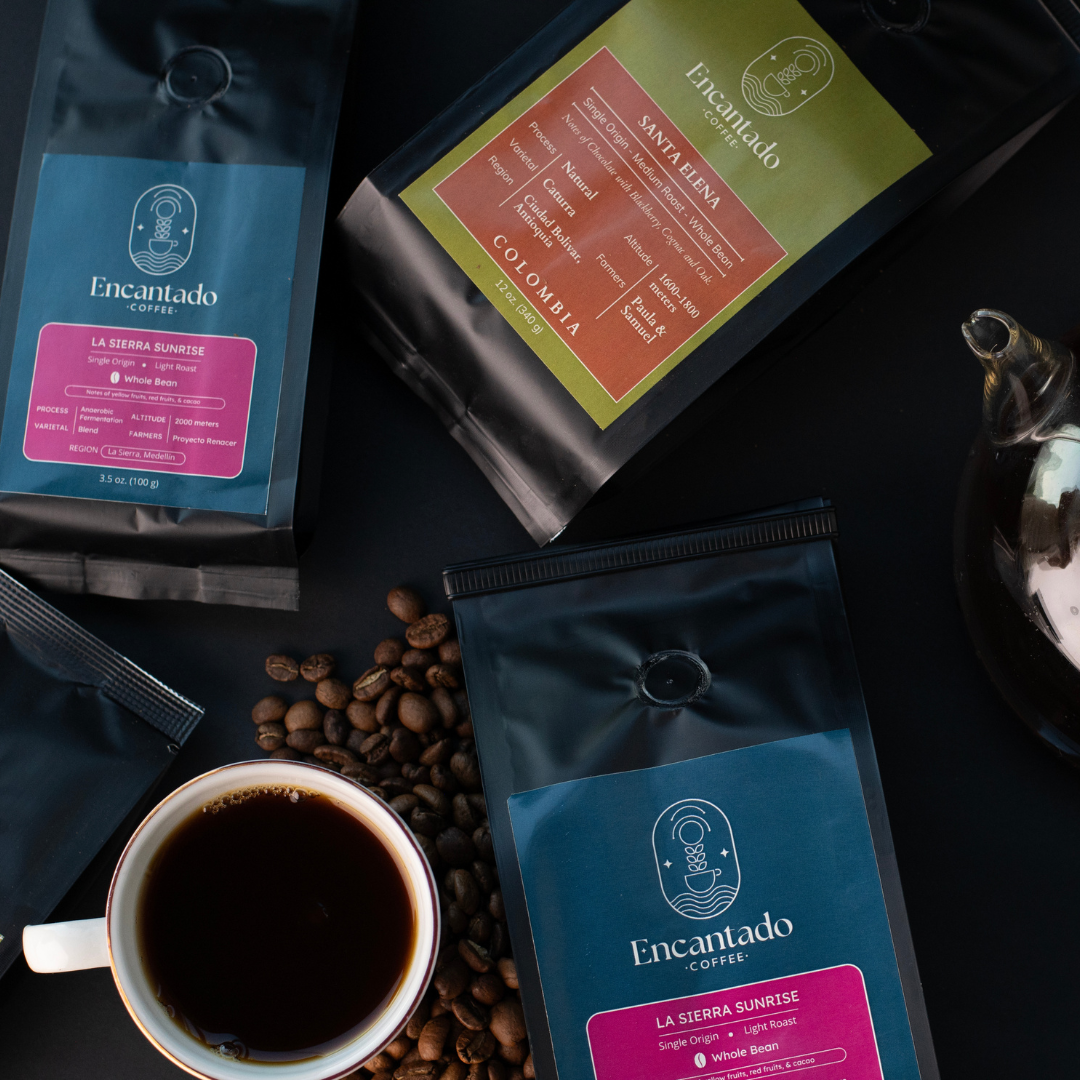 Three bags of coffee with blue and green labels sit on a black table next to a cup of coffee with scattered coffee beans